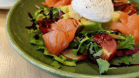 Close-up view of salad with salty salmon, avocado, poached egg and leaf vegetables on green plate in restaurant. Soft focus. Slow motion handheld video. Healthy eating theme.