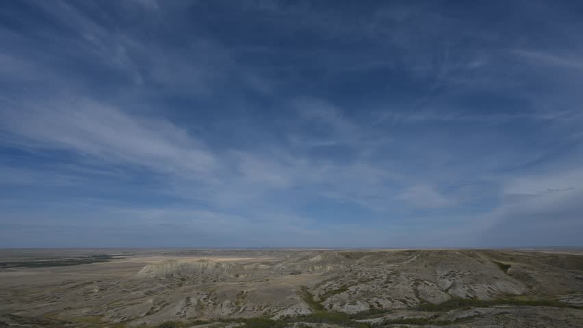 Time lapse of white wispy clouds in a blue sky above a hilly badlands desert setting. Scrubby trees occur in some valleys and a dirt road winds through the hills.  
