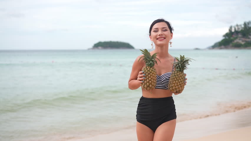 Asian tanned beautiful woman in black bikini with pineapple in her hands on beach tropical island background with sea ocean beach copy space. Happy girl smiling with organic fruits. Travel concept.