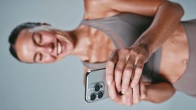 Vertical video studio portrait of smiling woman wearing fitness clothing against grey background messaging on mobile phone - shot in slow motion