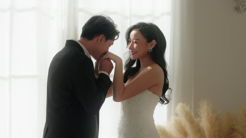 Captured in a soft glow, an Asian groom expresses his devotion with a gentle hand kiss to his bride on their wedding day, a perfect Valentine Day moment of love and respect, Love and Wedding Concept
