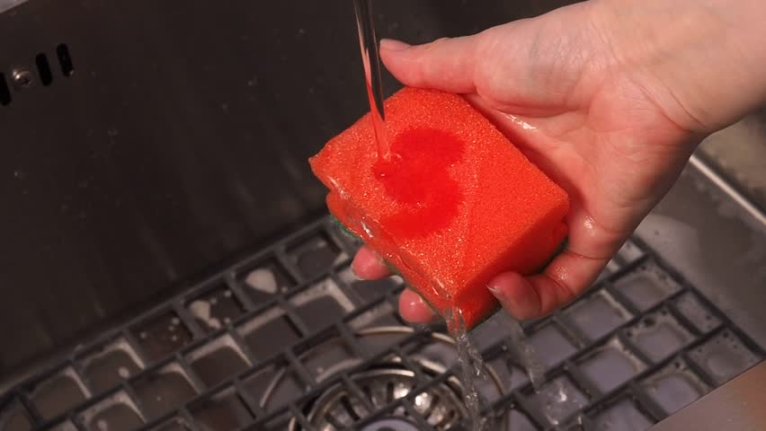 A sponge for washing dishes. The housewife holds a sponge in her hands. Water pours from the tap into the sink. Moisten the sponge with water. Royalty-Free Stock Footage #3393124277