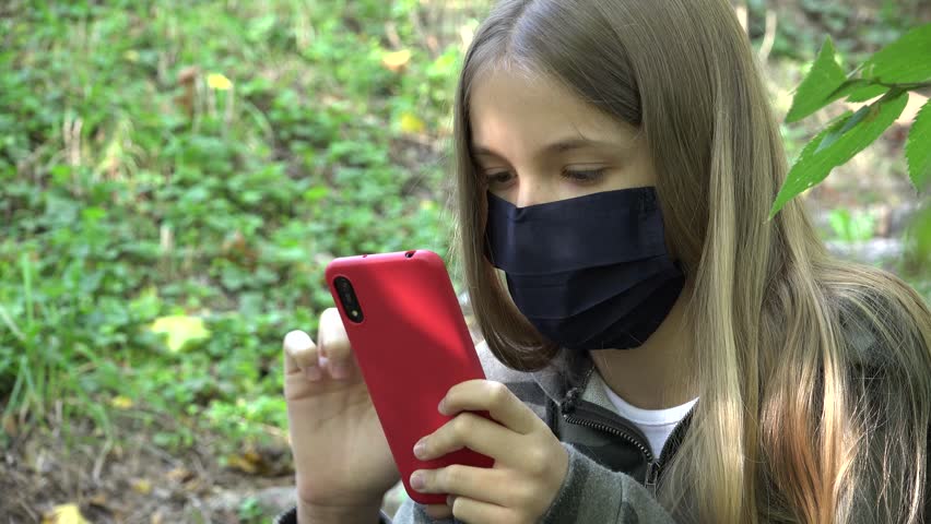 Sick Child with Protective Mask due Coronavirus Pandemic, Kid Browsing Internet on Smartphone in Park, Girl Playing Smart Phone