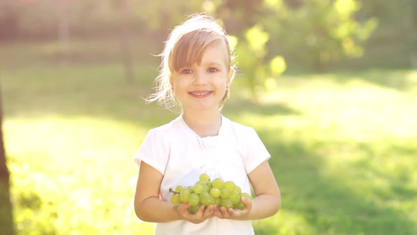 Girl holding grapes and looking at camera. Sunny day outdoors.

