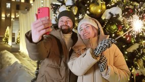 A couple of friends waving fireworks and filming greetings using a smartphone near an illuminated Christmas tree in the city center. A handsome man and a young woman in warm casual clothes take selfie