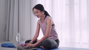 An Asian woman in workout attire uses her laptop to find exercise information on a yoga mat in her home's sitting room. Ideal for showcasing a healthy lifestyle and home yoga practices.