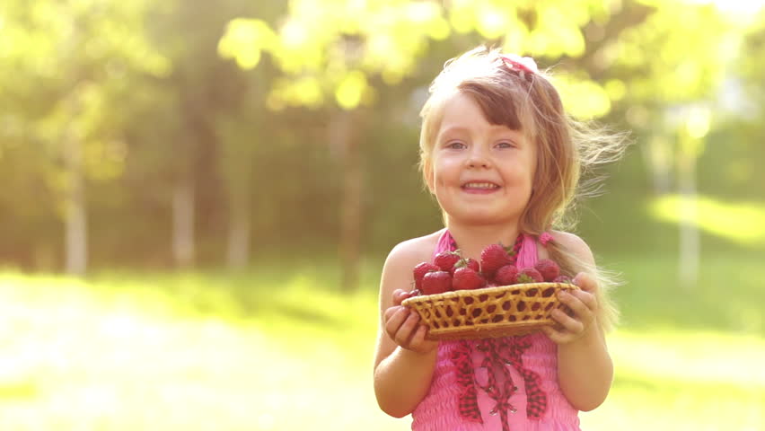 Child holding basket of strawberries and looking at camera in sunny day. Slow