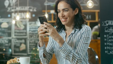 Beautiful Woman Sits in the Cafe Uses Smartphone. She Smiles while Messaging Her Friends or Loved One. On Her Table Cup with Beverage and Croissant. In the Background Busy Stylish Coffee House. 