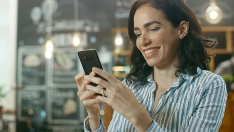 Beautiful Woman Sits in the Cafe Uses Smartphone. She Smiles while Messaging Her Friends or Loved One. In the Background Stylish Coffee House and Busy Waiter. Shot on RED EPIC-W 8K Helium Cinema Camer