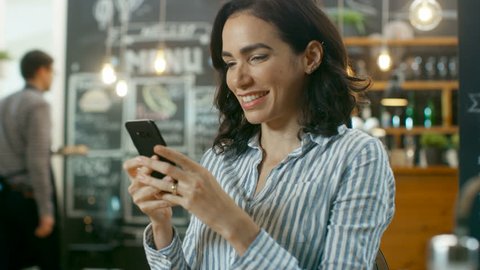 Beautiful Woman Sits in the Cafe Uses Smartphone. She Smiles while Messaging Her Friends or Loved One. In the Background Stylish Coffee House and Busy Waiter. Shot on RED EPIC-W 8K Helium Cinema Camer