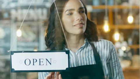 Beautiful Young Cafe Owner Turning Storefront Sign From Close to Open and Welcoming Customers into Stylish Coffee Shop. Big City with Traffic and People Visible in Window Reflection. 4K UHD.