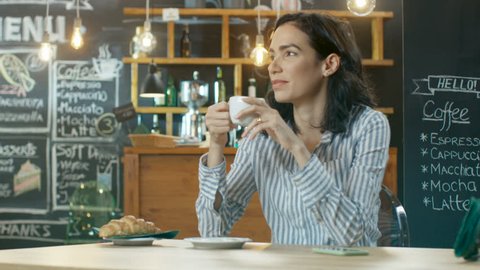 Beautiful Hispanic Woman Relaxes in Cafe, Drinking Beverage from the Cup, Looks at Her Mobile Phone where Incoming Message Bleeped. In the Background Stylish Modern Coffee House. Shot on RED EPIC-W 8K