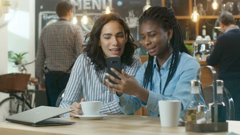 Two Beautiful Girlfriends Sitting in the Coffee Shop and Chatting, One Shows Pictures on Her Smartphone, they Both Laugh. Shot on RED EPIC-W 8K Helium Cinema Camera.