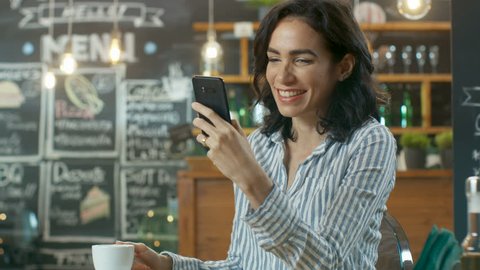 Beautiful Woman Sits in the Cafe Uses Smartphone. She Smiles while Browsing through Social Media. In the Background Busy Stylish Coffee House. Shot on RED EPIC-W 8K Helium Cinema Camera.