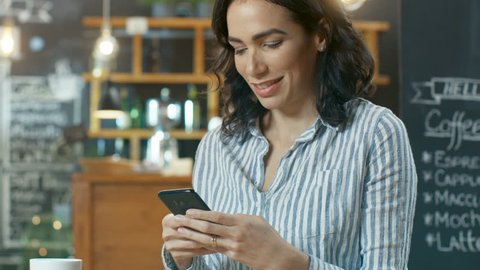 Beautiful Woman Uses Smartphone while Sitting in the Cafe. She Drinks Beverage from the Cup. Background of This Stylish Coffee Shop. Shot on RED EPIC-W 8K Helium Cinema Camera.