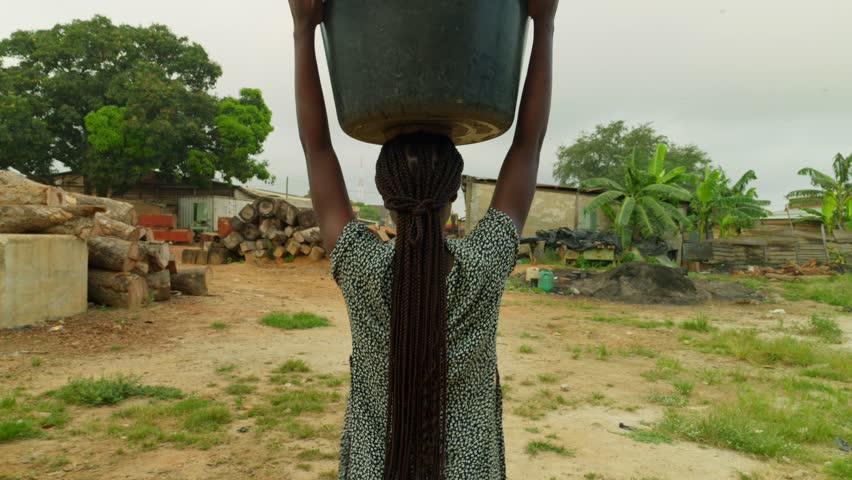After fetching some water, a village woman is carrying a heavy container with water on top of her head in her neighborhood in Kumasi, Ghana, in Africa. Royalty-Free Stock Footage #3393411689