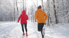 Middle-aged smiling trail couple runners man and woman dressed bright windproof jackets running by picturesque snowy forest path during sunny frosty day. Sporty active people, winter training 4K video