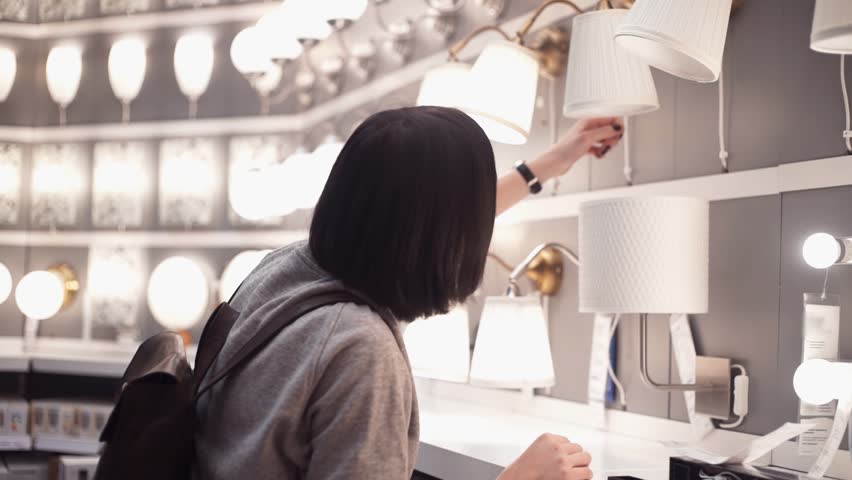 Customer chooses bedroom goods in hypermarket. Young woman examining light bulb in lamp in department store. Slow motion Royalty-Free Stock Footage #33935332