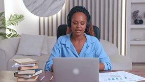 Young beautiful successful African American woman online teacher gesturing teaching students via internet talking about importance of productivity in work sits at table with books and laptop.