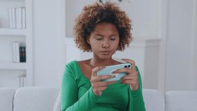 Young attractive ethnic African American woman teenager plays video game in smartphone and throws gadget on couch after losing dressed in casual clothes sits on white sofa inside house.