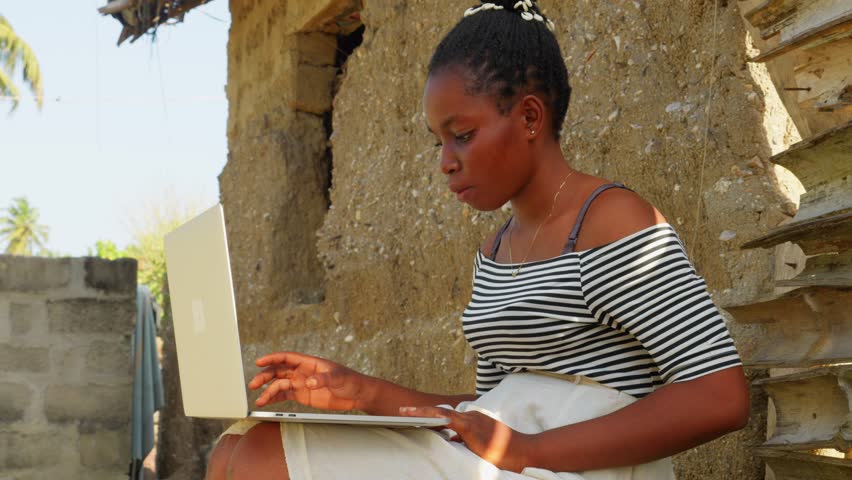 young black female girl working in her modern laptop while sitting in remote village in Africa , concept of technology mixed with lifestyle in africa poor country Royalty-Free Stock Footage #3393727611