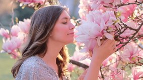 Spring beauty portrait of a beautiful young woman smells blooming magnolia flower. Spring blossom. Gorgeous romantic adult girl in dress near blooming magnolia tree outdoors