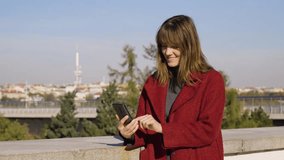 An attractive middle-aged Caucasian woman talks on a video call on a smartphone with a smile by a wall in an urban area - a cityscape in the blurry background