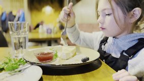 A little girl sits at a table in a restaurant with great pleasure and eats a fresh vanilla cheesecake with strawberry jam.