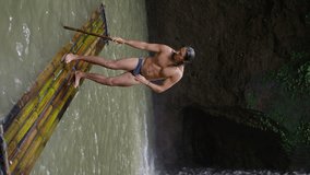 A man with long black hair and a beautiful torso floats on a wooden raft in a mountain river, slow motion, vertical view
