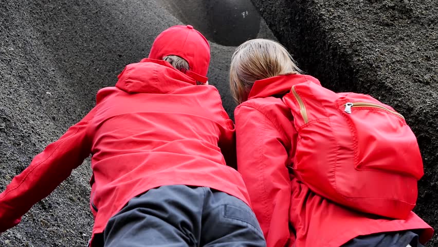 Looking from behind, two teenage hikers, a boy and a girl, stand close to a black rock and point their finger up the canyon. Hiking into the crater of a volcano.