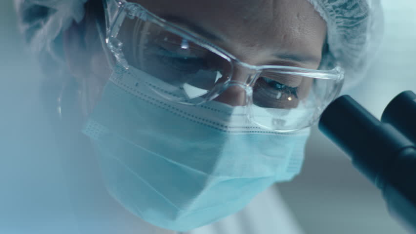 Close up shot of female lab tech wearing protective face mask, safety glasses and sterile hat using microscope and writing down notes during scientific research Royalty-Free Stock Footage #3393987163