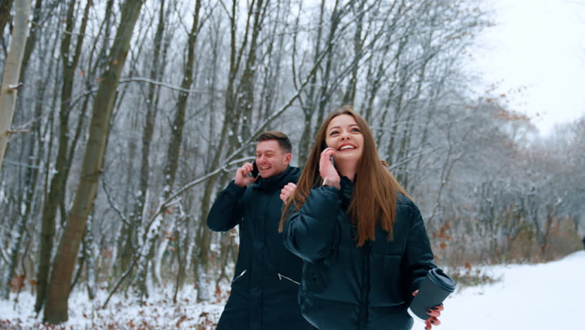 Smiling woman with paper cup in hand walks speaking on the phone. Man following behind laughs speaking on his phone too.