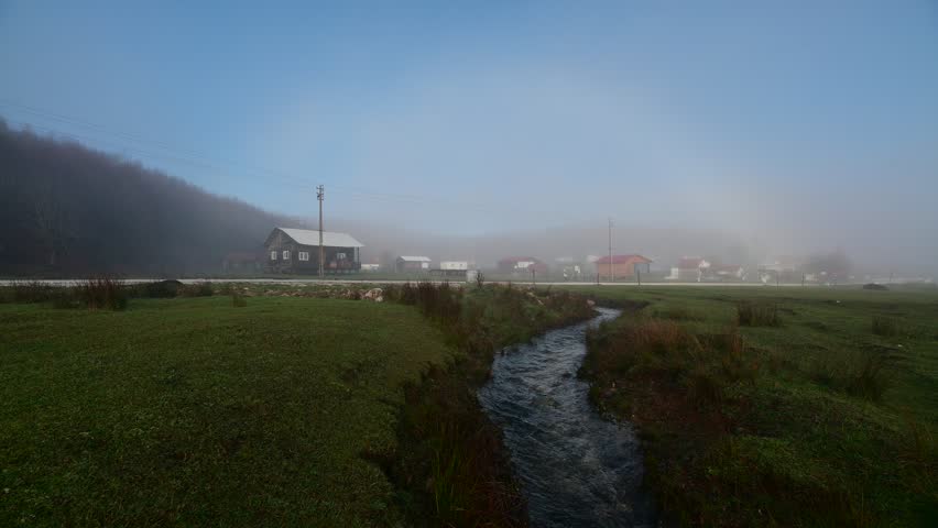 A narrow stream winds through a misty green plateau toward a distant rural building beneath low clouds. The wide highland view emphasizes wet grassland, fog, and quiet open terrain.