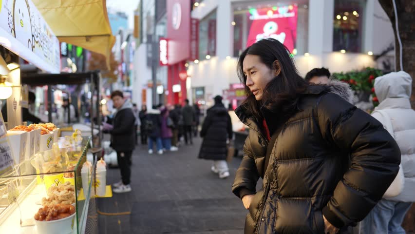 A South Korean man in his 30s with long hair, sightseeing at night in the winter snowfall on a street in Myeongdong, Seoul, South Korea.