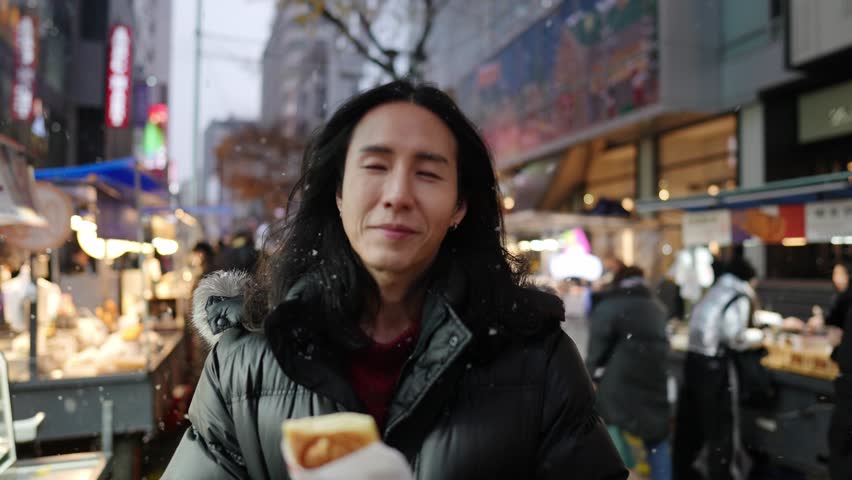 A South Korean man in his 30s with long hair, sightseeing at night in the winter snowfall on a street in Myeongdong, Seoul, South Korea.