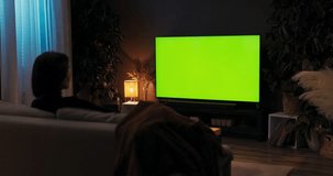 A young lady unwinding after a long day at work by viewing television while seated on a sofa in her comfortable living room, green screen mockup.