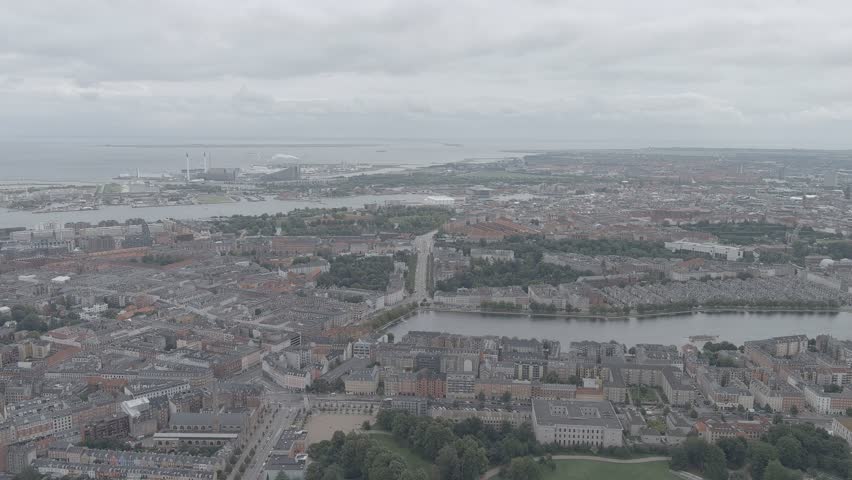 Copenhagen, Denmark. The Copenhagen Lakes curve through the foreground in a distinctive arc, forming a natural boundary between the dense historic city center and the surrounding districts. Beyond the lakes, the urban fabric stretches toward the harbor and port area, where industrial facilities and waterfront infrastructure meet the sea. In the far distance, a bridge spans the horizon, connecting the Danish coast. The scene is captured from a high aerial perspective on an overcast summer day, with diffused light softening the muted palette of gray rooftops, green parkland, and calm water surfaces. The slow forward motion of the camera reveals the layered depth of the city, from the tree-lined lakeside paths to the distant maritime skyline, conveying the scale and coastal character of the Danish capital. A-Log.