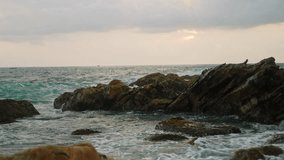 Sunrise over rocky shore, waves crash against boulders, seabirds perch atop. Tropical coastline with lighthouse overlooking turbulent sea. Serene seascape video for nature enthusiasts, travel vlogs.