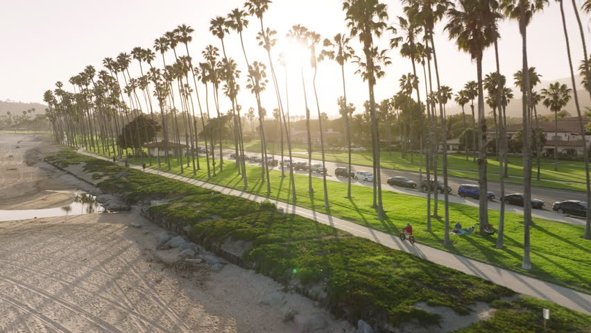 Panoramic view of tourists walking on road along Pacific coast of Santa Barbara, California, USA. Drone flying along city coastline with palm trees at sunset. City life at the ocean, 4k footage 