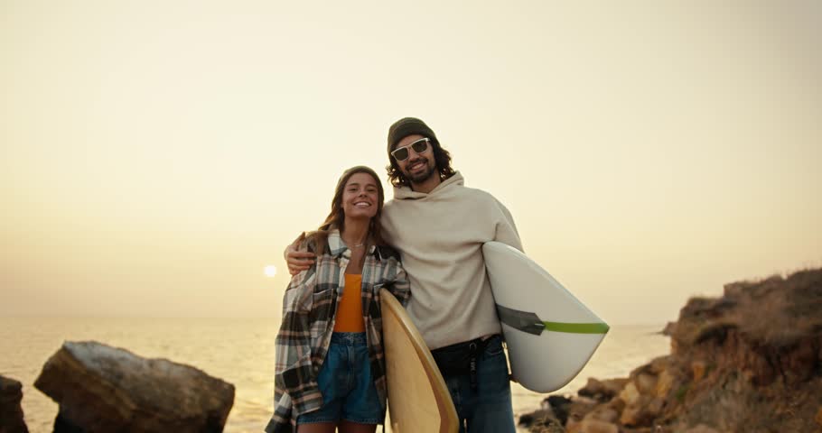 Portrait of a happy couple, a brunette man in a white sweatshirt and black sunglasses together with his blonde girlfriend in a plaid shirt stand near the sea and hold surfboards in their hands in the Royalty-Free Stock Footage #3394441547