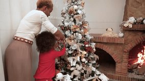 African Dark-skinned Mom and child hang a Christmas ball on a decorated Christmas tree next to a fireplace with fire and have fun. Family decorating the Christmas tree. Christmas time
