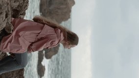 Vertical video. Young Lonely Woman Sits on Sharp Rocks in a Pink Raincoat, Gazing at the Sea with Waves and Rocks Behind Her.