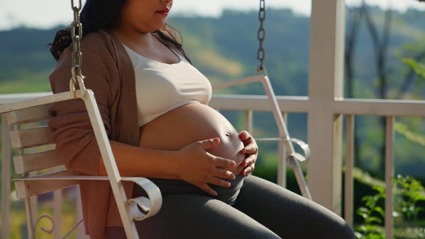 Winter of pregnant woman sitting on swing at a rural suburb house. Atmosphere is great, calm, and good weather, suitable for children's growth. Happy woman relaxes, enjoys rubbing her belly. Royalty-Free Stock Footage #3394573741