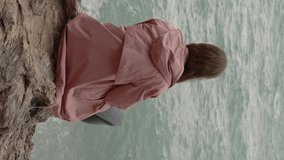 Vertical video. Lonely Woman Sits on Jagged Rocks and Contemplatively Watches the Strong Waves from the Storm in the Sea.