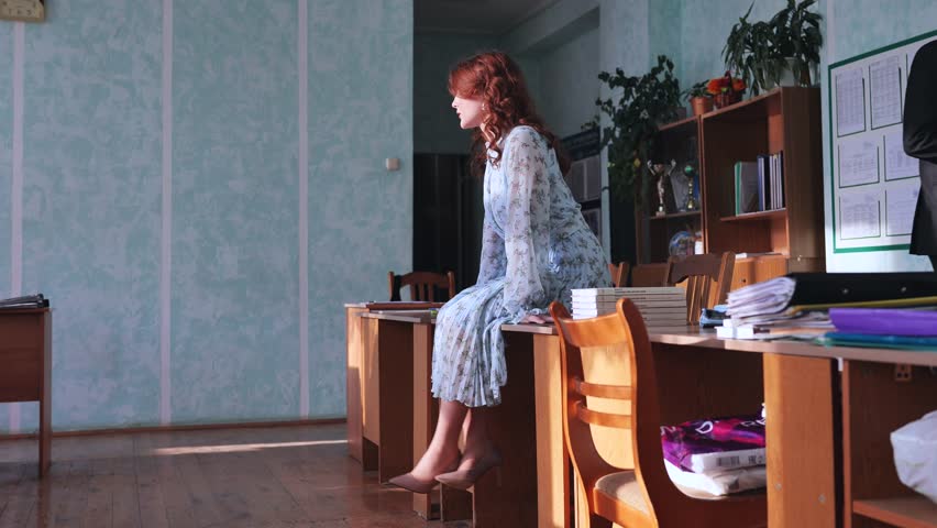 A beautiful schoolgirl in a dress and heels sits on a desk in class.