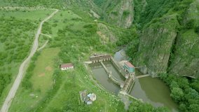 Aerial view of a hydroelectric power station on a river surrounded by high rocky cliffs in summer in cloudy weather, green grass, trees, village houses along the banks. Dzoraget River Canyon Armenia