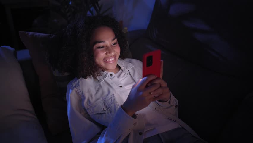 Joyful young black girl using cell phone lying on sofa. African woman enjoying chatting on mobile at night home. Single African American female generation z people on dating apps and social networks Royalty-Free Stock Footage #3394708403