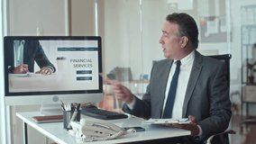 Medium shot of aged entrepreneur pointing at screen of computer with landing page of financial services and talking on camera