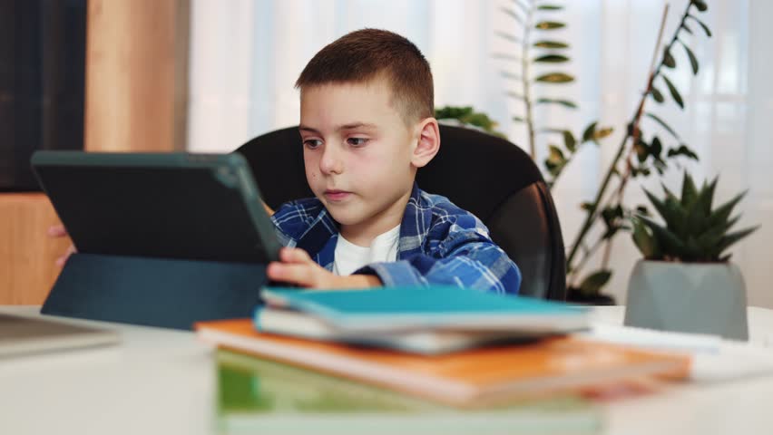 Clever young boy looking on tablet screen with focused facial expression while sitting at home desk with books and notes. Caucasian elementary pupil using digital device for distance education. Royalty-Free Stock Footage #3395008343