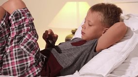 Closeup video of cute blond kid laying on back in bed at home interior. Child holds modern smart phone and playes cheerfully videogame. Real time full hd video footage.